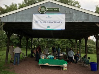 A picture of a pavilion with a sign on it that says Wildlife Sanctuary. Underneath the pavilion is a group of people conversing.